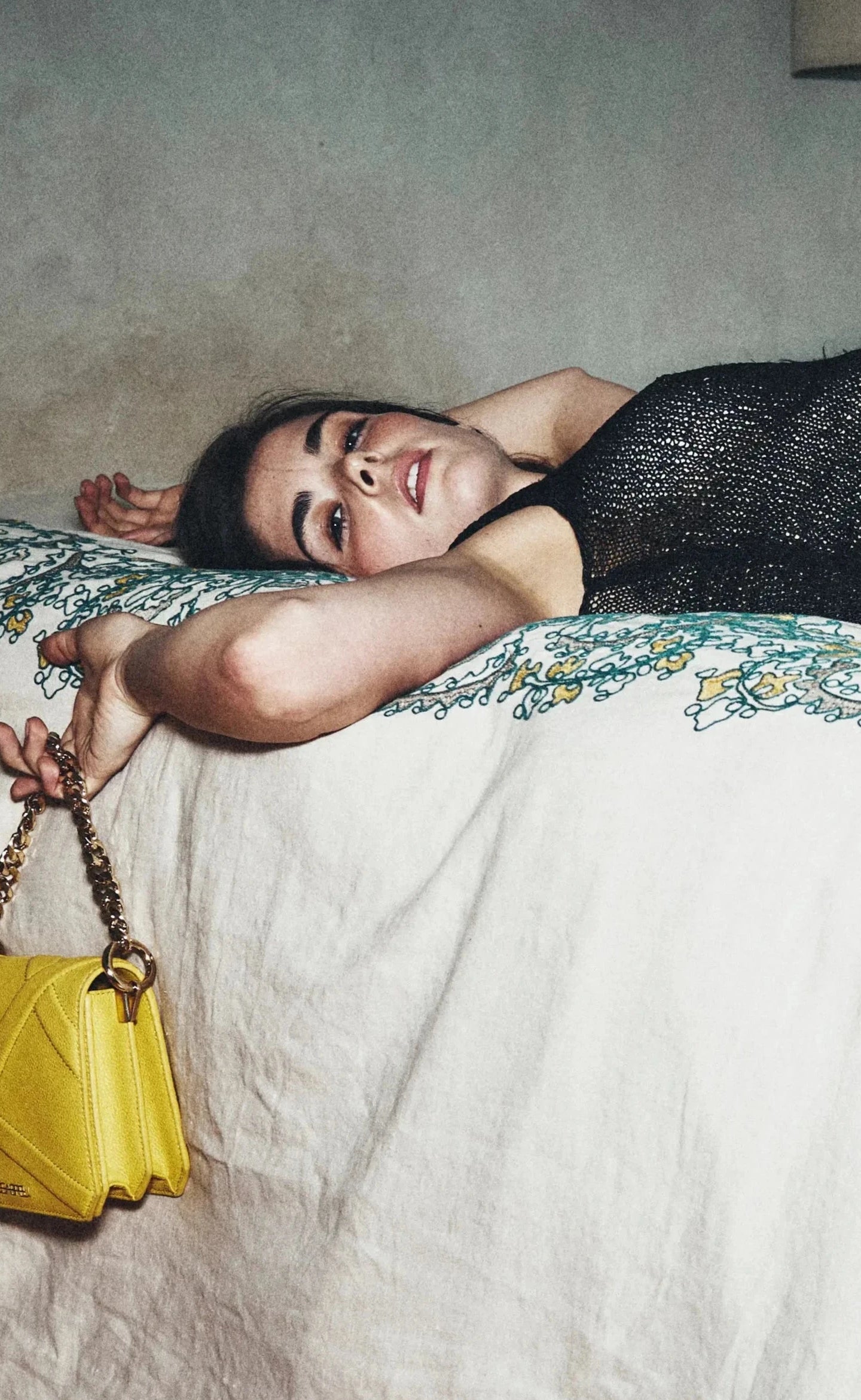Woman lying on a bed wearing a black dress and stockings, holding a yellow handbag.